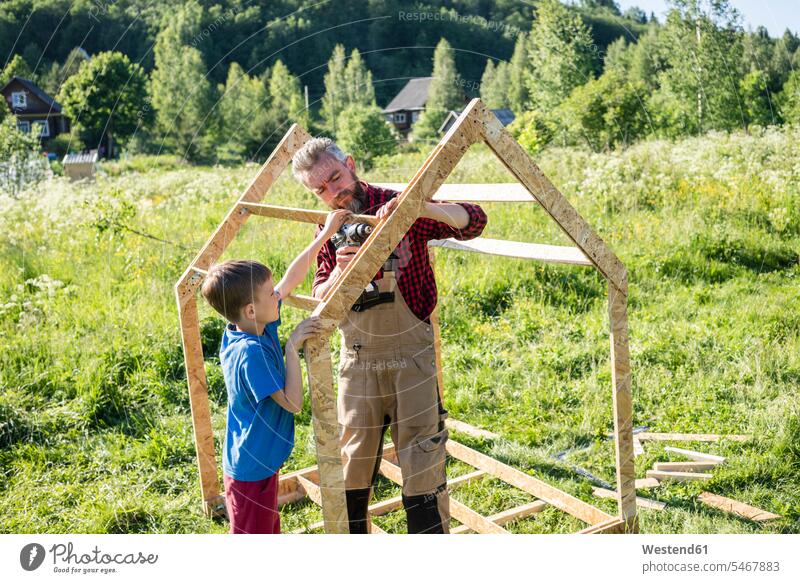 Junge hilft Zimmermann beim Bau eines Spielhauses an einem sonnigen Tag Farbaufnahme Farbe Farbfoto Farbphoto Außenaufnahme außen draußen im Freien