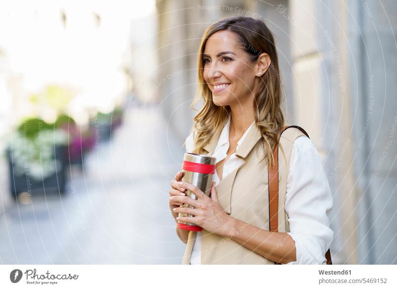 Lächelnde Frau hält metallene Thermoskanne, während sie am Bürgersteig in der Stadt steht Farbaufnahme Farbe Farbfoto Farbphoto Außenaufnahme außen draußen