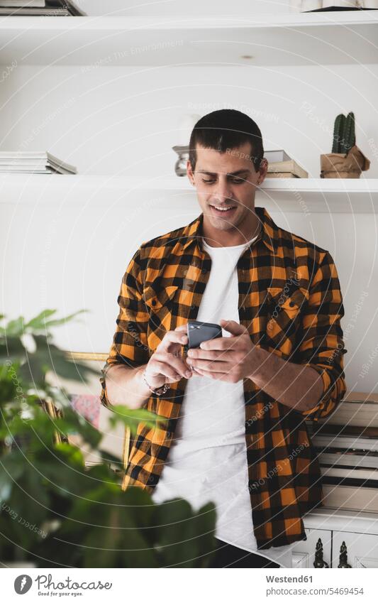 Junger Mann steht an einem Regal und benutzt ein Mobiltelefon Männer männlich Ablage Regale Handy Handies Handys Mobiltelefone stehen stehend Erwachsener