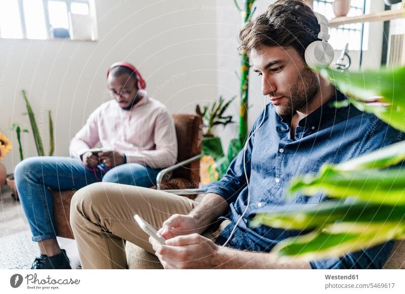 Zwei junge Männer mit Handys und Kopfhörern sitzen in Sesseln in einem Loft Mobiltelefon Handies Mobiltelefone Lofts Mann männlich Kopfhoerer sitzend sitzt