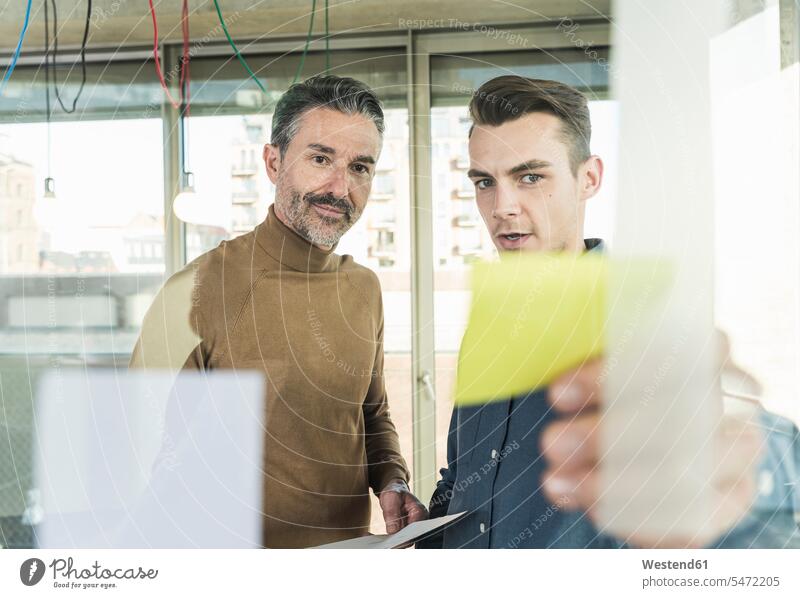 Reifer Geschäftsmann und junger Mann arbeiten an Haftnotizen an einer Glasscheibe im Büro Arbeitskollege Arbeitskollegen Kollege Partner Partnerschaften Job