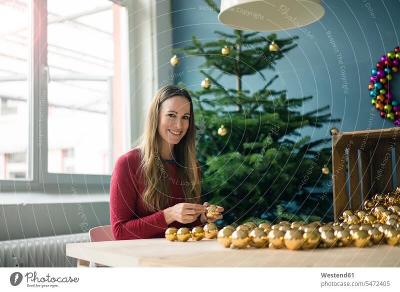 Porträt einer lächelnden Frau am Tisch sitzend mit vielen goldenen Weihnachtskugeln goldfarben goldfarbene Christbaumkugel Christbaumkugeln Portrait Porträts