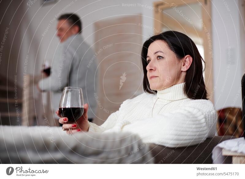 Seriöse reife Frau mit einem Glas Wein und Mann im Hintergrund Trinkgläser Gläser Trinkglas Wohnzimmer Wohnraum Wohnung Wohnen Wohnräume Wohnungen Rotwein