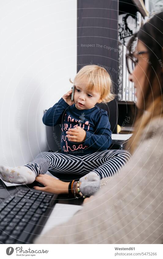 Mutter arbeitet zu Hause mit kleinen Tochter sitzt auf dem Schreibtisch hält Handy sitzen sitzend Arbeitstisch Schreibtische Zuhause daheim Mobiltelefon Handies