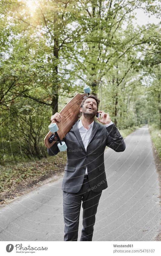 Glücklicher Geschäftsmann mit Skateboard spricht mit Smartphone auf Landstraße Rollbretter Skateboards Businessmann Businessmänner Geschäftsmänner Landstraßen