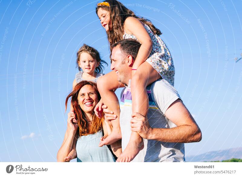 Fröhliche Eltern, die ihre Töchter bei Sonnenschein auf den Schultern gegen den klaren Himmel tragen Farbaufnahme Farbe Farbfoto Farbphoto Spanien