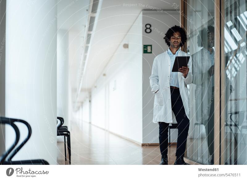Selbstbewusster junger männlicher Arzt, der ein digitales Tablet benutzt, während er sich an eine Glaswand im Krankenhauskorridor lehnt Farbaufnahme Farbe
