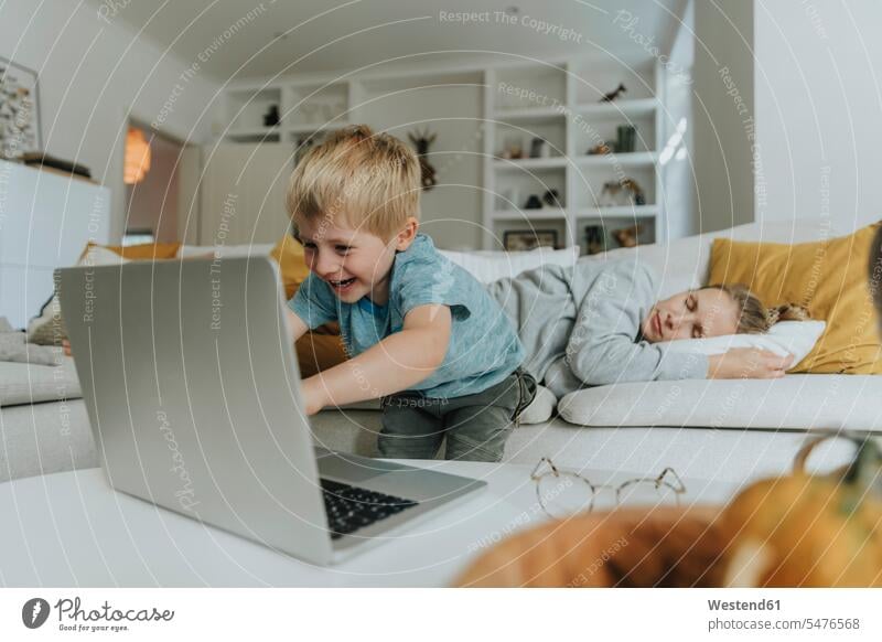 Junge benutzt Laptop der Mutter, die zu Hause auf dem Sofa schläft Farbaufnahme Farbe Farbfoto Farbphoto Innenaufnahme Innenaufnahmen innen drinnen Tag