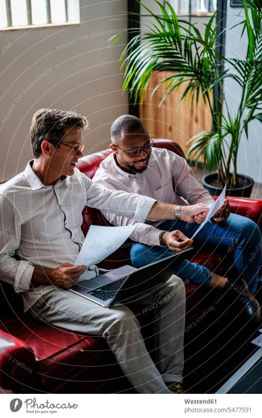 Zwei Geschäftsleute mit Laptop und diskutieren Dokumente auf Sofa in Loft-Büro Office Büros besprechen Besprechung Couches Liege Sofas Papiere Unterlagen