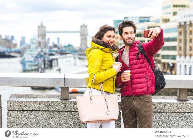 UK, London, junges Paar mit Kaffee zum Mitnehmen steht auf der Brücke über die Themse und macht ein Selfie mit Smartphone Pärchen Paare Partnerschaft iPhone