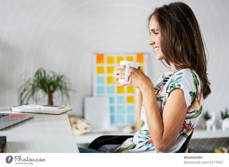 Lächelnde junge Frau trinkt Kaffee am Schreibtisch Arbeitstisch Schreibtische trinken lächeln weiblich Frauen Tisch Tische Erwachsener erwachsen Mensch Menschen