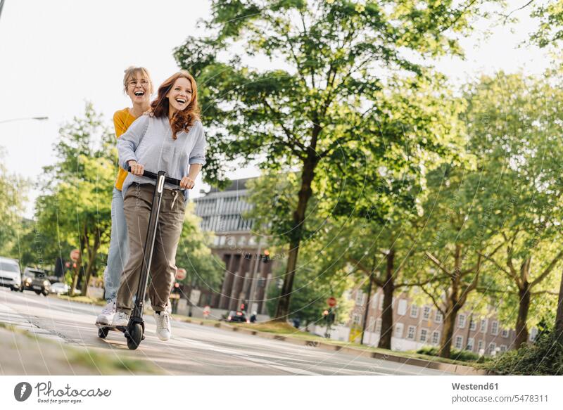 Glückliche Freundinnen fahren gemeinsam E-Scooter in der Stadt Freunde Kameradschaft begeistert Enthusiasmus enthusiastisch Überschwang Überschwenglichkeit