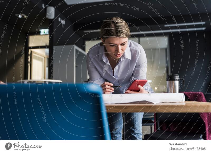 Junge Frau mit Handy und Plan am Tisch im Büro Mobiltelefon Handies Handys Mobiltelefone weiblich Frauen Pläne Office Büros Tische Telefon telefonieren