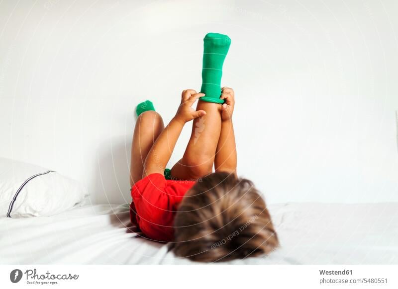 Rückenansicht eines kleinen Jungen, der auf dem Bett liegt und seine grünen Socken anzieht Betten Buben Knabe Knaben männlich Kind Kinder Kids Mensch Menschen