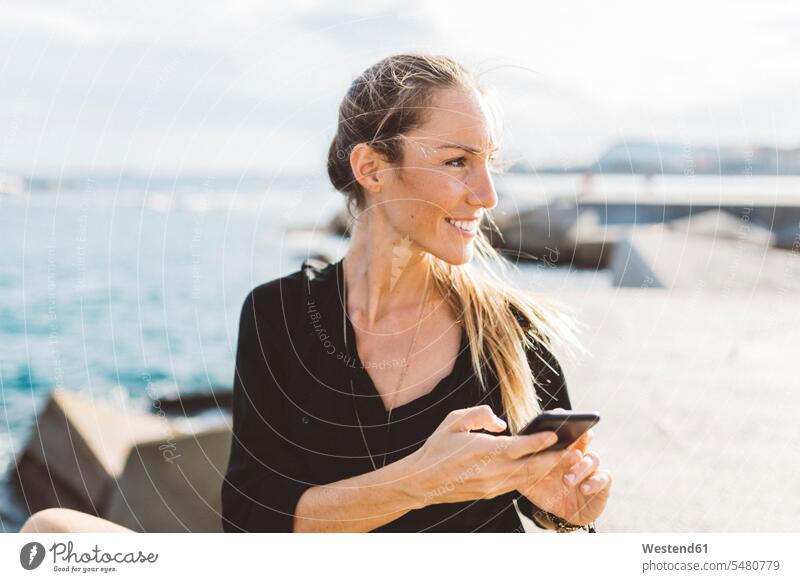 Lächelnde junge Frau mit Handy an der Strandpromenade, die sich umsieht weiblich Frauen Mobiltelefon Handies Handys Mobiltelefone lächeln glücklich Glück