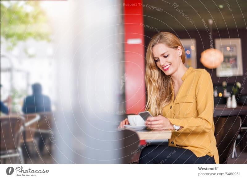 Lächelnde junge Frau sitzt in einem Café und schaut auf ein Smartphone weiblich Frauen Cafe Kaffeehaus Bistro Cafes Cafés Kaffeehäuser iPhone Smartphones