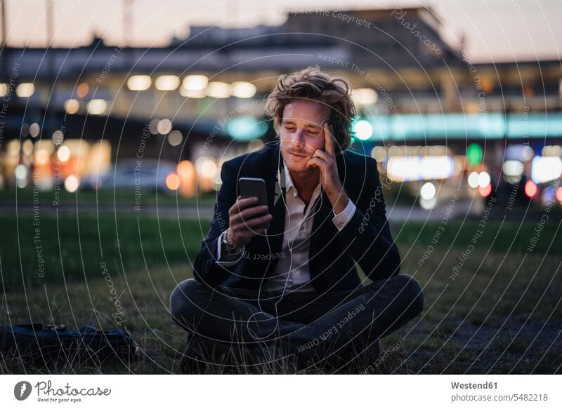 Geschäftsmann sitzt in der Dämmerung mit Handy und Kopfhörern auf der Wiese Businessmann Businessmänner Geschäftsmänner Mobiltelefon Handies Handys