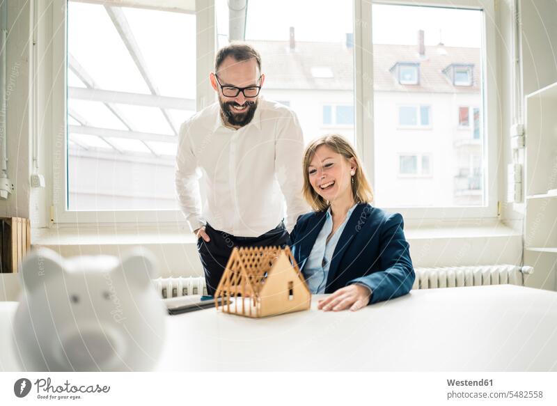 Lachende Frau und lachender Mann mit Hausmodell und Sparschwein im Amt Sparschweine Geschäftsfrau Geschäftsfrauen Businesswomen Businessfrauen Businesswoman