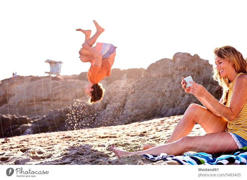 Junger Mann am Strand macht einen Salto mit einer Frau, die ihr Handy kontrolliert Saltos Salti Mobiltelefon Handies Handys Mobiltelefone Paar Pärchen Paare