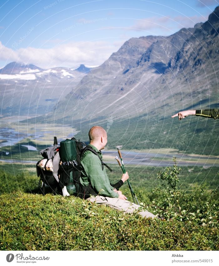 Eine Hand zeigt auf einen Wanderer der im Profil mit Rucksack an einem grünen Hang sitzt und den Ausblick in ein Tal genießt Ausflug Abenteuer Berge u. Gebirge