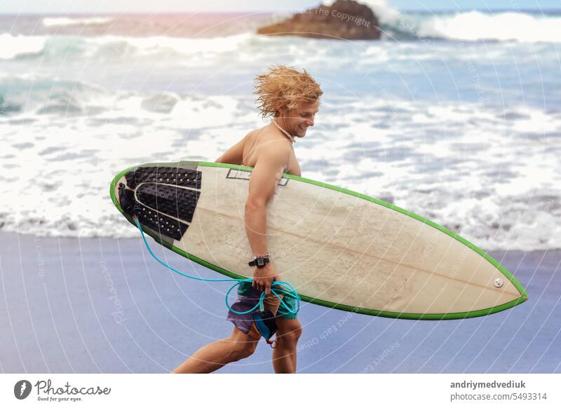 Fit jungen Surfer Mann mit lockigem blondem Haar mit Surfbrett geht durch den Ozean Spaß haben extreme Wassersportarten, Surfen. Reisen und gesunden Lebensstil Konzept. Sport Reiseziel.