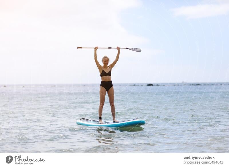 Glückliche junge Frau in stilvollen schwarzen Bikini hob das Paddel, Balancieren auf einem SUP-Board, auf ruhigen Ozean Wasser auf sonnigen Sommertag. Gesunder Lebensstil und extreme Erholung Konzept.