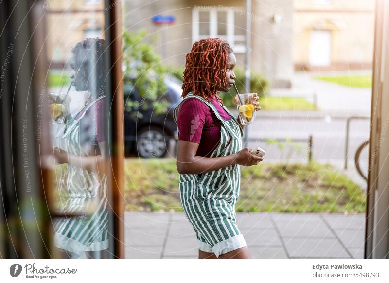 Junge nicht-binäre Person, die in der Tür eines Cafés steht und Saft trinkt echte Menschen positiv selbstbewusst Erwachsener junger Erwachsener Lifestyle