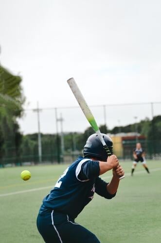 Softballspielerin / Baseballspieler mit dem Rücken zur Kamera hat einen heranfliegenden Ball fest fixiert und holt aus zum Schlag um ihn zu treffen Rasen