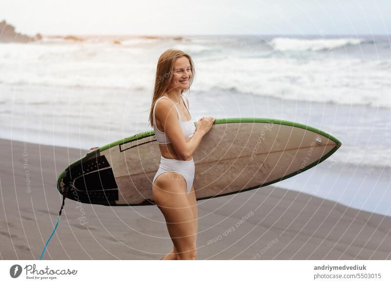 Junge Surferin im Bikini geht surfen steht mit Surfbrett auf dem schwarzen Sandstrand. Mädchen geht mit Longboard. Extreme Wassersport am Meer, Meer. Gesunder aktiver Lebensstil. Sommer-Urlaub