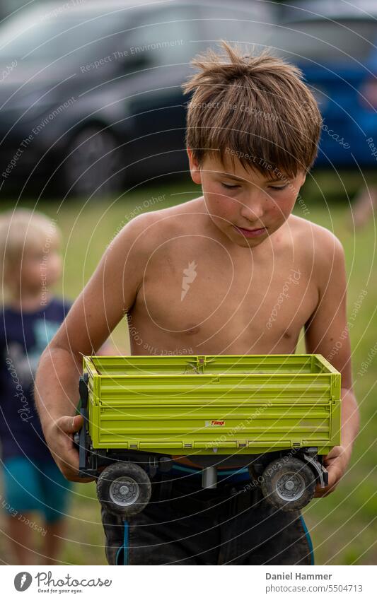10 jähriger Junge trägt einen Anhänger von einem Spielzeugtraktor. Er ist konzentriert und freut sich auf das Spielen mit seinen Freunden. Im Hintergrund ein kleinerer Junge, der nicht erkennbar ist und eine grüne Wiese mit parkenden unscharfen Autos.