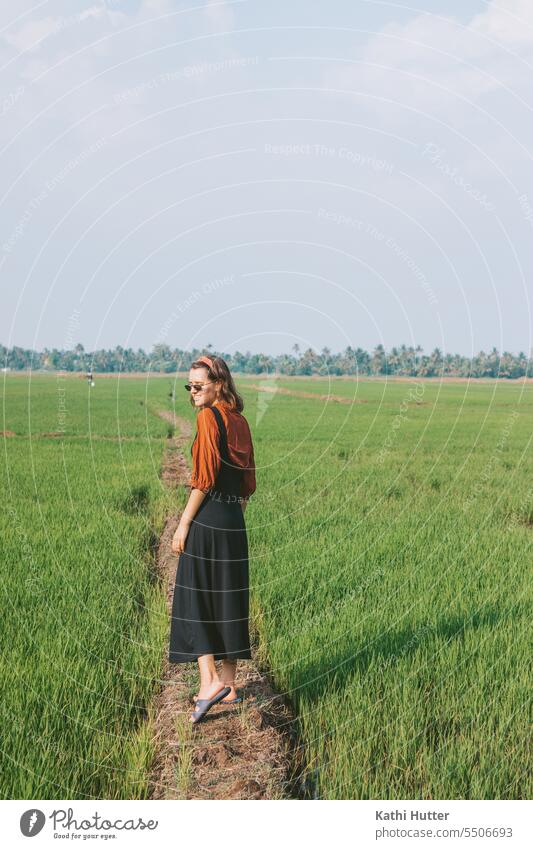 eine junge Frau ist auf einem Reisfeld in Kerala, Indien. Sie trägt ein schwarzes Kelid mit einer roten Bluse darunter. Sonnenbrille Landschaft grün Asien Natur