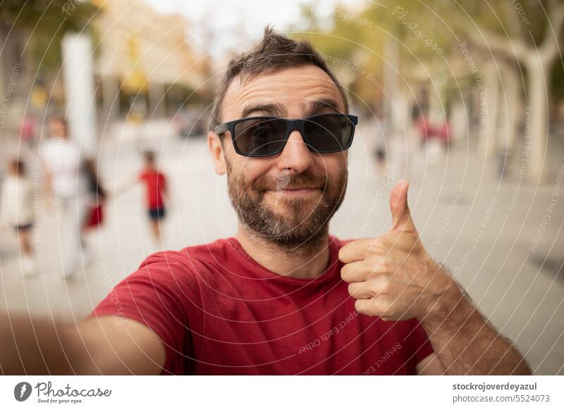 Ein junger, bärtiger Mann mit Sonnenbrille lächelt in die Kamera, während er bei einem Spaziergang in der Stadt ein Selfie mit seinem Handy macht. Person Männer