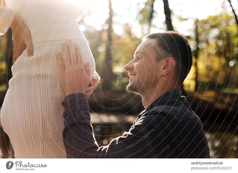 Cropped Schuss von glücklichen jungen Mann genießt Schlaganfälle, Gespräche und küsst Bauch der Frau, warten auf Geburt Baby im Freien in Sonnenstrahlen im Stadtpark. Mutterschaft pränatale Versorgung und Frau Schwangerschaft Konzept.