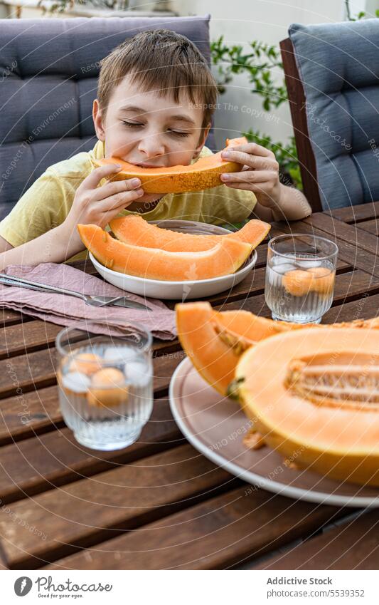 Preteen boy sitting at table and eating fresh cut musk melon Junge Kind essen Frucht frisch Gesundheit Melonen Scheibe Natur Sommer Eis Erfrischung trinken