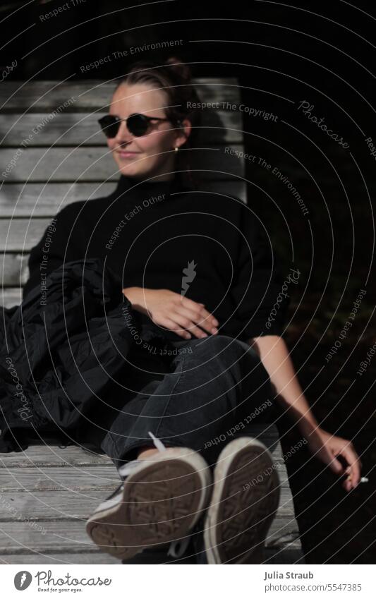 Schöne Frau genießt eine Zigarette auf der Bank Pause machen Holzbank Füsse oben liegen schwarz gekleidet Sonnenbrille jung Chucks Herbst