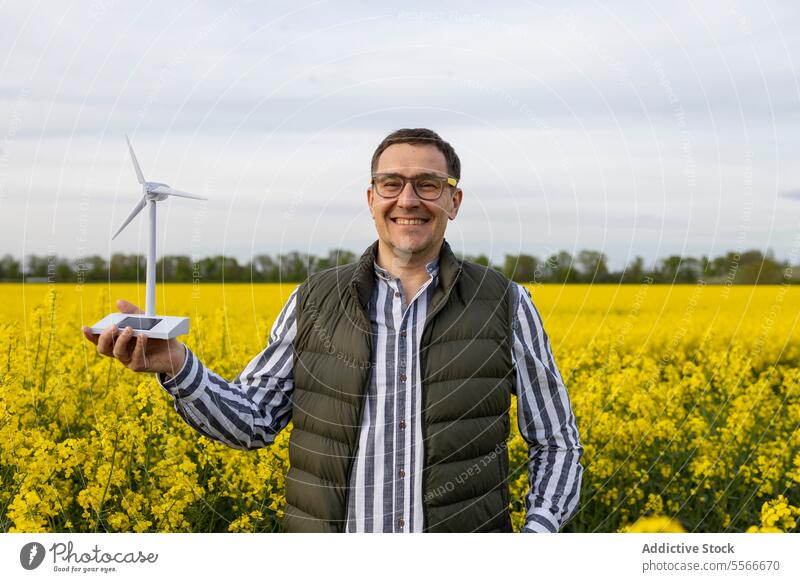 Älterer Mann mit Brille, der ein Windradmodell auf einer blühenden Wiese präsentiert. Windkraftanlage Model Feld Blume gelb präsentieren Lächeln Natur stehen