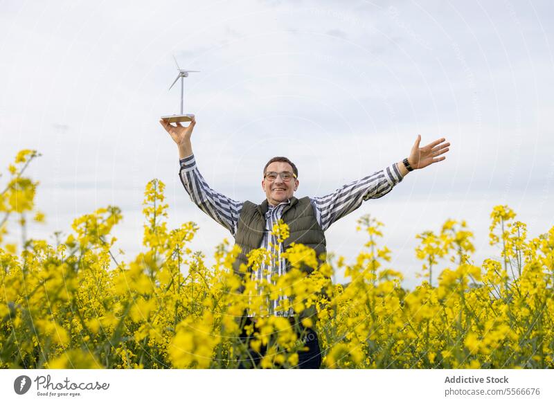 Fröhlicher reifer Mann mit Brille, der inmitten von Blüten erneuerbare Energie feiert. Windkraftanlage Model Feld Blume gelb anheben Freude Waffen Feier Natur