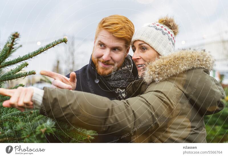 Paar, das einen Weihnachtsbaum kaufen möchte und mit dem Finger auf den ausgewählten Baum zeigt Zeigefinger Familie skeptisch Rotschopf Tradition Wahl Lächeln