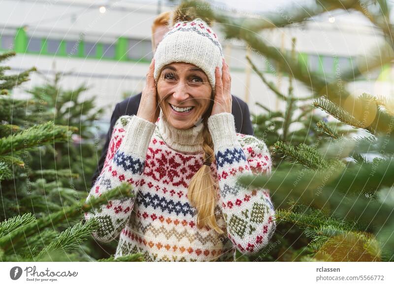 Glückliche Frau und ihr Freund wollen auf einem Markt einen Weihnachtsbaum kaufen und schauen in die Kamera überrascht Familie skeptisch Rotschopf Tradition