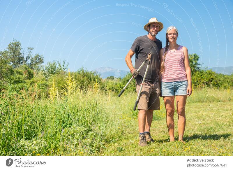 Reifes Paar mit Werkzeug auf offenem Feld stehend Mann Frau Hut Stroh Werkzeuge Gartenarbeit gestreift Tank Top Berge Hintergrund im Freien Natur sonnig Stehen