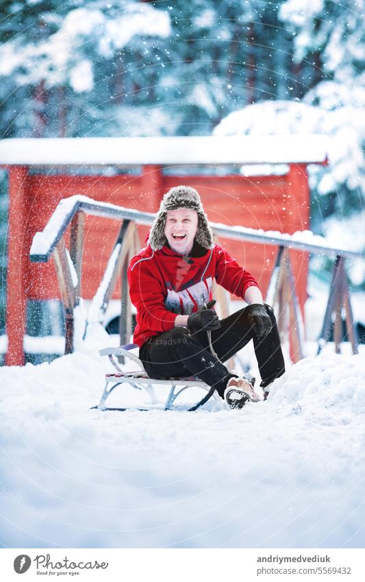Lächelnde junge fröhliche kaukasischen Mann trägt Wurm roten Sweatshirt und Spaß Winter Ohr Hut hat Spaß mit Schlitten in einem verschneiten Park oder Wald am Wintertag. Frohe Weihnachten Winterurlaub