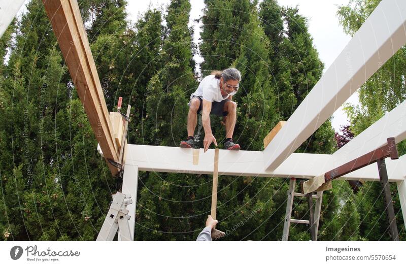 Balance. Männlicher Arbeiter steht auf Holzbalkenkonstruktion für Carport. Übergabe des Hammers auf der Baustelle. Arbeitssicherheitskonzept stehend