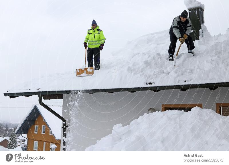 Zwei Männer schaufeln hohen, schweren Schnee von einem Hausdach schneeräumen männer schneien winter schneeschaufeln haus hausdach schneelast gewicht gefahr
