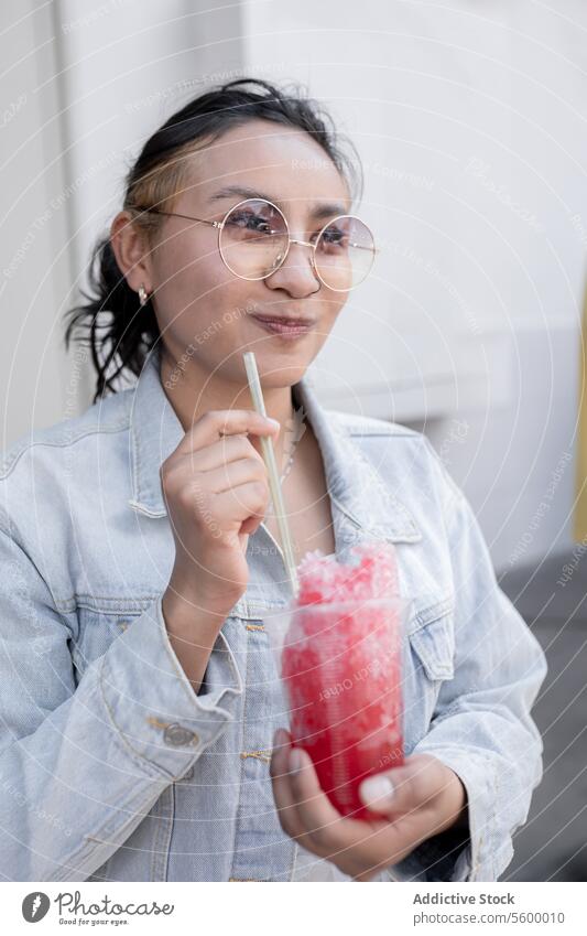 Eine junge hispanische Frau trinkt lächelnd einen mexikanischen Kirschslushy im Freien Sonnenbrille Schwarzes Haar blond Highlights Ohrringe Jeansstoff Jacke