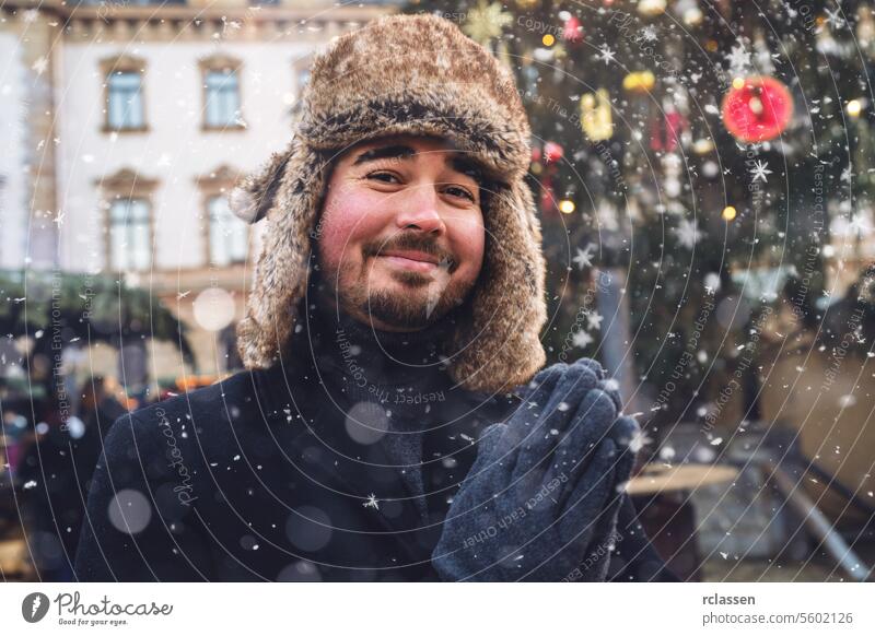Mann in Pelzmütze und Wintermantel lächelt sanft, Schneeflocken fallen um ihn herum, Weihnachtsmarktbeleuchtung im Hintergrund. Frohe Weihnachten Tasse Kakao
