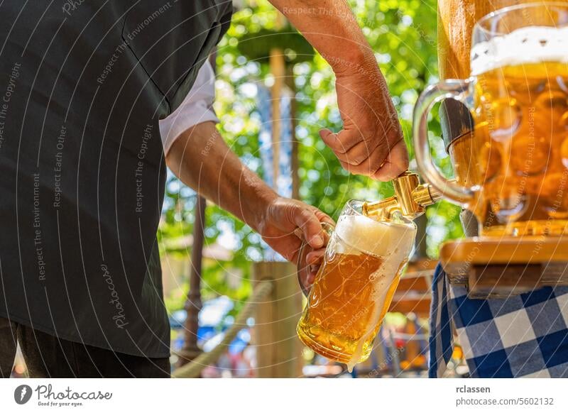 Bayerischer Mann in Schürze schenkt ein großes Lagerbier aus einem Holzfass im Biergarten ein. Hintergrund für Oktoberfest oder Wiesn, Volks- oder Bierfest