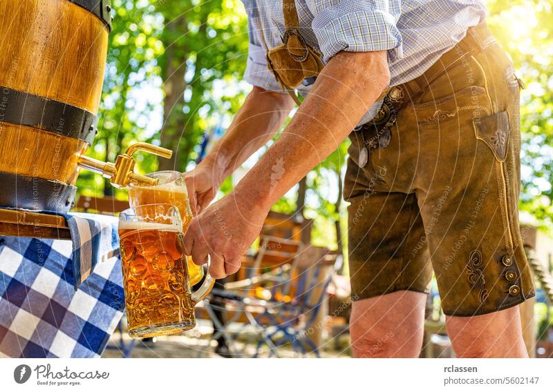 Ein bayerischer Mann in traditioneller Lederhose zapft im Biergarten ein großes Lagerbier aus einem hölzernen Bierfass. Hintergrund für Oktoberfest oder Wiesn, Volks- oder Bierfest