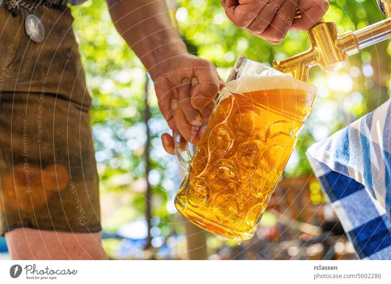 Ein bayerischer Mann in Lederhosen schenkt ein großes Lagerbier aus einem hölzernen Bierfass im Biergarten ein. Hintergrund für Oktoberfest oder Wiesn, Volks- oder Bierfest (deutsch: O'zapft is!)