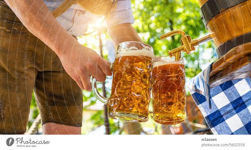 Ein bayerischer Mann in Lederhosen schenkt im Biergarten ein großes Lagerbier aus einem hölzernen Bierfass aus. Hintergrund für Oktoberfest oder Wiesn, Volks- oder Bierfest (deutsch: O'zapft is!)
