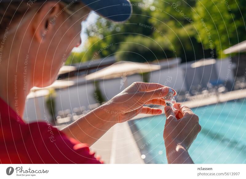 Nahaufnahme von Frauenhänden, die ein kleines Fläschchen für einen PH-Wert-Test schließen, mit Schwimmbad im Hintergrund Abfüllung Prüfung von Schwimmbadwasser